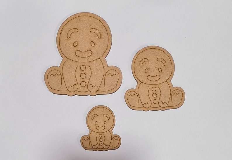 May include: Three unfinished wooden gingerbread man ornaments of varying sizes. The gingerbread men are sitting with their arms crossed and have a simple smiling face.
