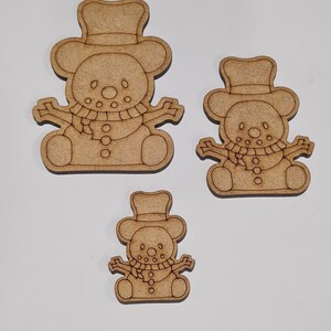 May include: Three wooden snowman decorations of varying sizes. Each snowman has a hat, scarf, and buttons. The decorations are unfinished and ready to be painted.