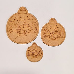 May include: Three wooden Christmas baubles with a winter village scene inside. The ornaments are different sizes and have a brown wood finish.