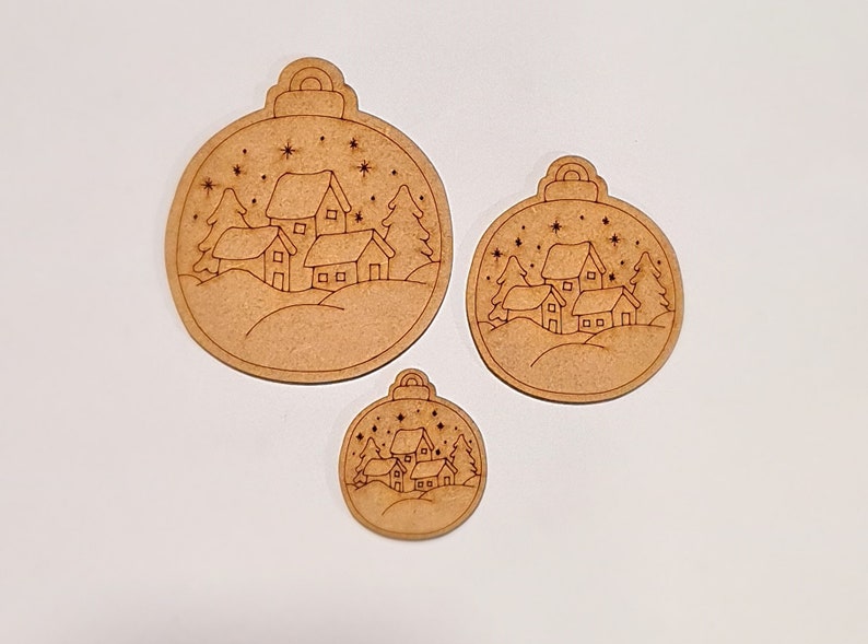 May include: Three wooden Christmas baubles with a winter village scene inside. The ornaments are different sizes and have a brown wood finish.