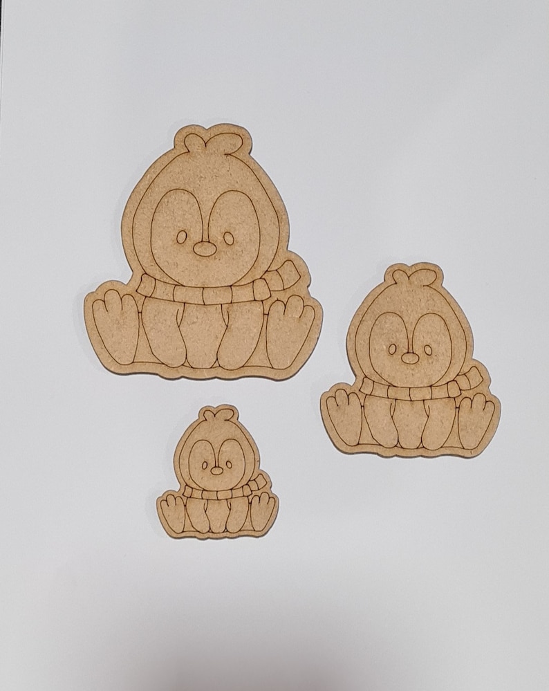 May include: Three unfinished wooden penguin ornaments of varying sizes. The penguins are wearing scarves and have simple facial features.