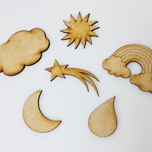 May include: Six wooden cutouts of weather elements: a sun, a cloud, a crescent moon, a raindrop, a shooting star, and a rainbow with clouds. The cutouts are all made of unfinished wood and have a natural brown colour.