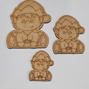May include: Three unfinished wooden elf ornaments in various sizes. The elves are wearing pointy hats and have big eyes and smiles. They are holding small bags in their hands.