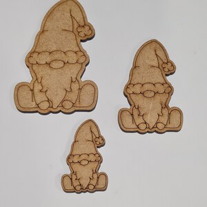 May include: Three wooden gnome ornaments in various sizes. The gnomes are sitting with their hands on their laps and wearing a hat with a pom-pom.
