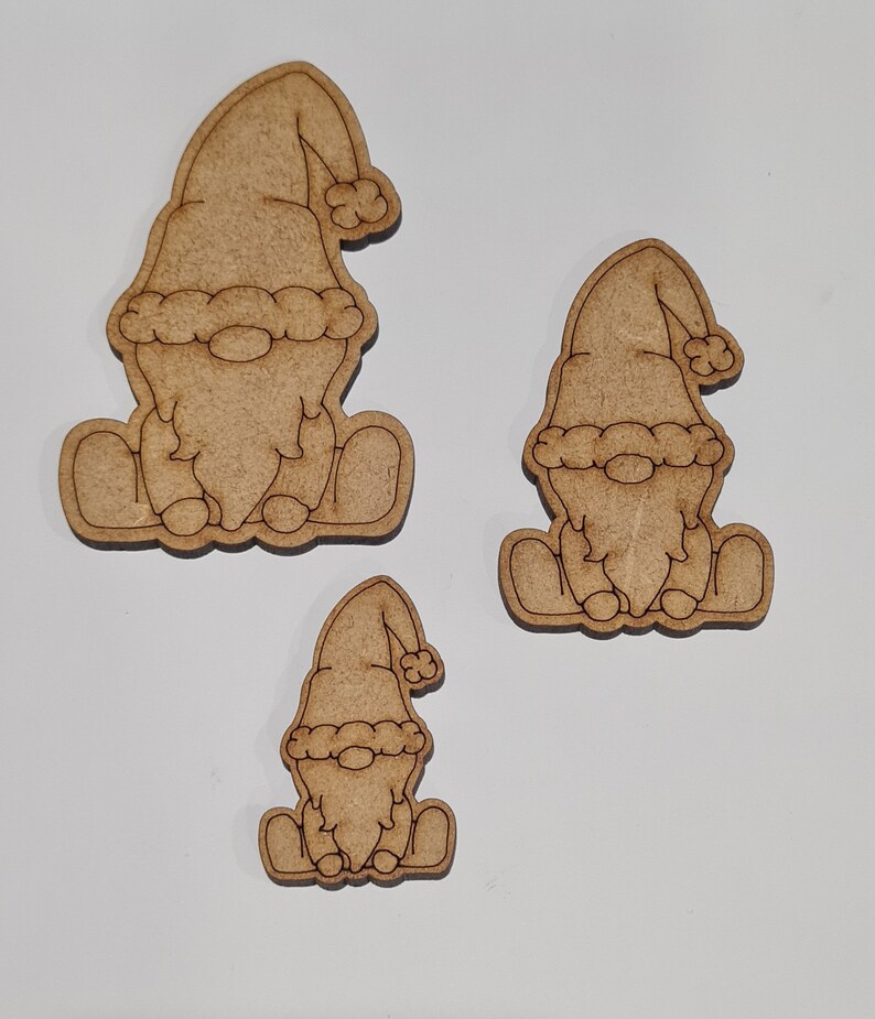 May include: Three wooden gnome ornaments in various sizes. The gnomes are sitting with their hands on their laps and wearing a hat with a pom-pom.