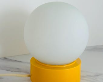 May include: A retro-style table lamp featuring a spherical, frosted white glass shade atop a bright yellow cylindrical base. The lamp has a simple design with a visible power cord. The lamp is on a marble surface.