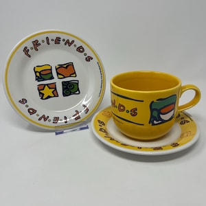 May include: A yellow ceramic cup and saucer set with the text "N.D.S" and a cartoon graphic. A matching plate with the word "FRIENDS" and colourful squares is also present. The set is on a white surface.