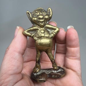 May include: A small, brass figurine of a smiling, elf-like character with large ears and arms akimbo. The figure has a rounded belly and stands on a small, rectangular base. The brass has a warm, aged patina.