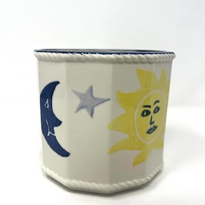 May include: White ceramic planter with a blue rim and rope-like trim. The planter features a whimsical design with a yellow sun, a blue crescent moon, and a grey star. The planter has a geometric shape.