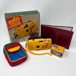 May include: A Big Yellow Camera gift set including a yellow 35mm camera with a wrist strap, a multi-coloured fabric carrying case in red, blue, and green, a maroon photo album, a box of Kodak Ultra Gold film, and the original cardboard packaging.