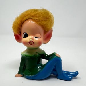 May include: A vintage ceramic elf figurine winking. The elf has a peach-coloured face, large ears, and yellow hair. It wears a green top and blue trousers. The figurine is in a seated position.