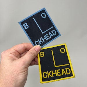 May include: Two square embroidered patches, each featuring the word "BLOCKHEAD". One has a black background with light blue trim and lettering. The other has a black background with yellow trim and lettering. The patches are held in a hand.