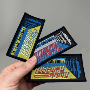 May include: Three rectangular embroidered patches with the text "TUBEWAY ARMY" in a retro font. The patches feature a black border, with yellow, blue, and white designs. The patches are held in a hand.