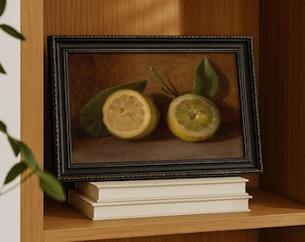 May include: A framed still life painting of two lemon halves with green leaves. The painting has a dark, ornate frame and rests on a stack of white books. The artwork is displayed on a wooden shelf, creating a classic and elegant aesthetic.