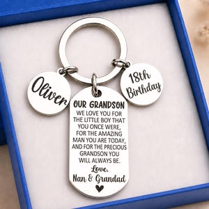 May include: A silver-toned metal keychain featuring a large rectangular tag engraved with the text OUR GRANDSON WE LOVE YOU FOR THE LITTLE BOY THAT YOU ONCE WERE, FOR THE AMAZING MAN YOU ARE TODAY, AND FOR THE PRECIOUS GRANDSON YOU WILL ALWAYS BE. Love, Nan & Grandad, plus two circular tags engraved with Oliver and 18th Birthday.