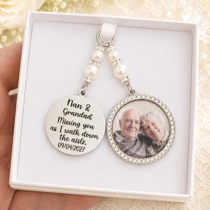 May include: A silver-toned memorial charm featuring two circular pendants hanging from white pearl beads. One pendant displays the text: Nan & Grandad Missing you as I walk down the aisle. 09/04/2027. The other pendant contains a photograph of an elderly couple.
