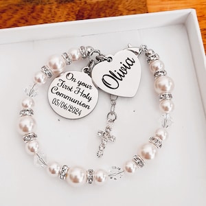 May include: A silver bracelet with white pearls and crystal accents. It features a heart charm with the name "Olivia" engraved on it and a round charm with the text "On your First Holy Communion 05/06/2024". A small silver cross charm hangs from the bracelet.