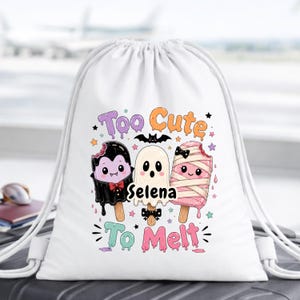 May include: A white drawstring backpack featuring a graphic of three cartoon popsicles: a vampire, a ghost, and a mummy. The text reads "Too Cute To Melt" with the name "Selena" in the centre, surrounded by small colourful stars and a black bat.