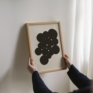 Puede incluir: Una l&aacute;mina de arte mural rectangular en un marco de madera clara. La obra presenta un grupo de c&iacute;rculos negros s&oacute;lidos de distintos tama&ntilde;os dispuestos en una formaci&oacute;n org&aacute;nica abstracta sobre un fondo de color blanco roto.