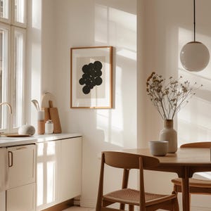 Puede incluir: Una l&aacute;mina de arte mural enmarcada que presenta un grupo de c&iacute;rculos negros sobre un fondo beige claro, expuesta en una cocina con una mesa de comedor de madera, sillas con asientos trenzados, una l&aacute;mpara colgante blanca y un jarr&oacute;n con flores secas.
