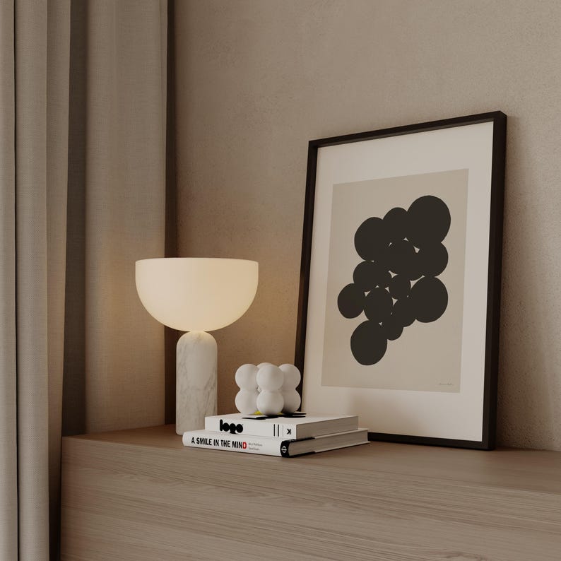 Puede incluir: Una l&aacute;mpara de mesa con base de m&aacute;rmol blanco y pantalla en forma de cuenco, una escultura blanca de esferas y dos libros apilados titulados Logo y A Smile in the Mind, junto a un cuadro con un conjunto de c&iacute;rculos negros sobre fondo beige.