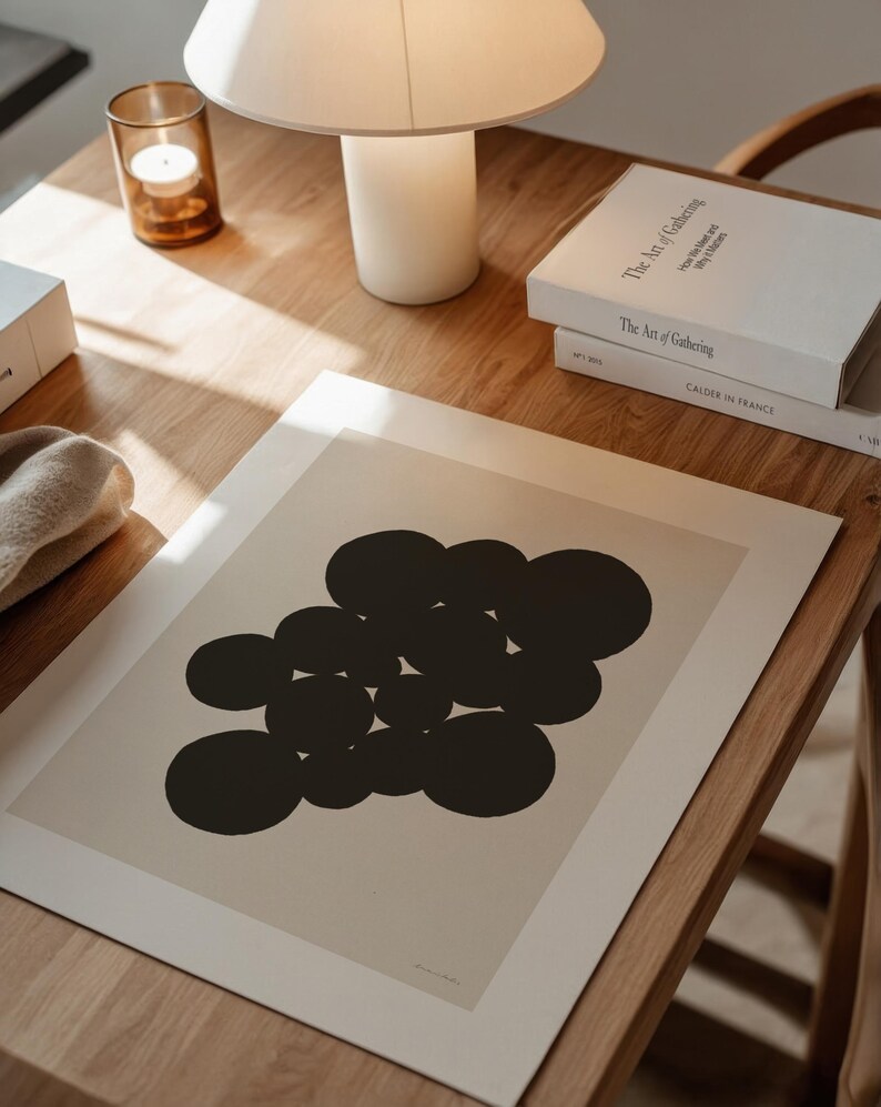 Puede incluir: Una l&aacute;mina de arte rectangular que presenta un grupo de c&iacute;rculos negros s&oacute;lidos dispuestos en un patr&oacute;n abstracto sobre un fondo beige claro. La l&aacute;mina se muestra sobre una superficie de madera junto a una l&aacute;mpara blanca, un portavelas y dos libros.