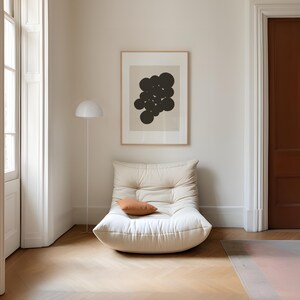 Puede incluir: Un sill&oacute;n bajo de color crema con capiton&eacute; y un peque&ntilde;o coj&iacute;n rectangular de color terracota. Sobre el sill&oacute;n cuelga una l&aacute;mina enmarcada con formas circulares de color marr&oacute;n oscuro sobre un fondo beige claro, junto a una l&aacute;mpara de pie blanca.