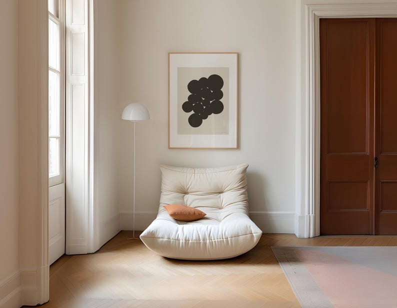 Puede incluir: Un sill&oacute;n bajo de color crema con capiton&eacute; y un peque&ntilde;o coj&iacute;n rectangular de color terracota. Sobre el sill&oacute;n cuelga una l&aacute;mina enmarcada con formas circulares de color marr&oacute;n oscuro sobre un fondo beige claro, junto a una l&aacute;mpara de pie blanca.