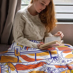 May include: A woman sits on a couch reading a book while wrapped in a colorful blanket featuring a graphic design of the Statue of Liberty, a cityscape, and the words "Avenue" and "Way".