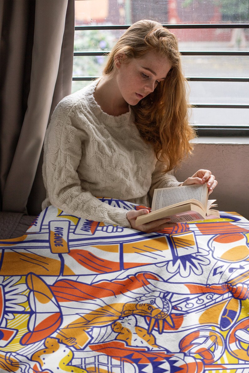 May include: A woman sits on a couch reading a book while wrapped in a colorful blanket featuring a graphic design of the Statue of Liberty, a cityscape, and the words "Avenue" and "Way".