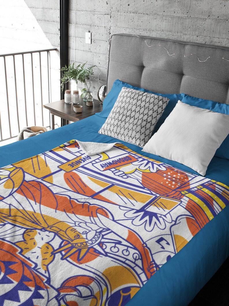 May include: A white fleece throw blanket with a colorful graphic design featuring a stylized image of the Statue of Liberty, a yellow taxi cab, and the text "BROADWAY" and "AVENUE".