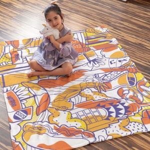 May include: A white and orange throw blanket with a colorful cartoon design featuring a hot dog, a phone booth, a slice of pizza, and the Statue of Liberty. The blanket is on a wooden floor and a young girl is sitting on it.