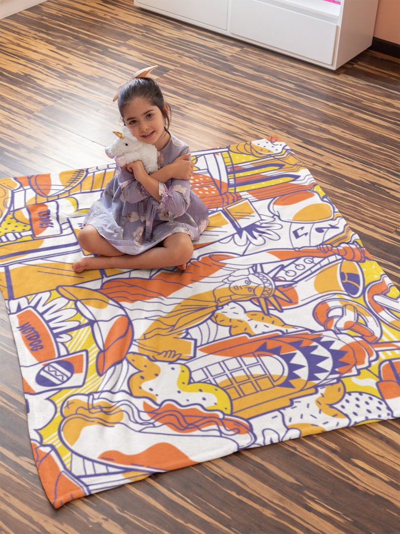 May include: A white and orange throw blanket with a colorful cartoon design featuring a hot dog, a phone booth, a slice of pizza, and the Statue of Liberty. The blanket is on a wooden floor and a young girl is sitting on it.