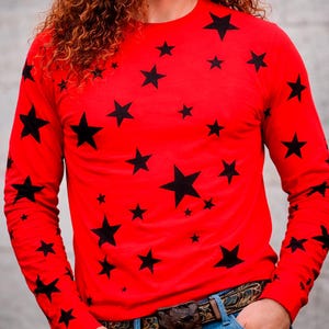 Könnte beinhalten: Ein rotes Langarmshirt mit einem Muster aus schwarzen Sternen. Die Sterne sind unterschiedlich groß und über das Shirt verteilt. Das Shirt hat einen Rundhalsausschnitt und ist aus weichem Material.