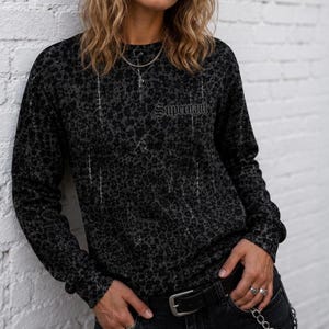May include: A long-sleeve crew neck shirt in a dark charcoal grey with an all-over black floral pattern. The shirt features the word Supernaut printed in a gothic-style font on the upper left chest area. It is paired with a black leather belt.