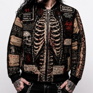Peut inclure: Un blouson bomber noir orné d'un imprimé graphique détaillé représentant une cage thoracique et une colonne vertébrale humaine. Le motif inclut divers textes comme Autopsy Division, des symboles de danger biologique et des étiquettes de dossier.