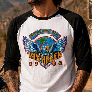 Puede incluir: Camiseta de béisbol blanca y negra con un gráfico colorido. El gráfico presenta un águila azul con las alas extendidas, un globo terráqueo y el texto "TOUR OF THE WORLD" y "VAN HALEN 1984".