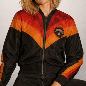 May include: A black bomber jacket featuring a chevron pattern in shades of red and orange across the chest and sleeves. The jacket has a front zip closure, ribbed cuffs, and a circular patch on the left chest with the text SUPERNAUT and a star.