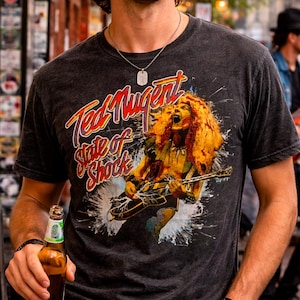Könnte beinhalten: Dunkelgraues T-Shirt mit einer Grafik von Ted Nugent, der Gitarre spielt, und dem Text "Ted Nugent State of Shock" in Rot und Gelb. Das Bild hat einen verwaschenen, Vintage-Look.