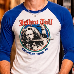 Può includere: T-shirt bianca e blu in stile raglan con la band Jethro Tull. La maglietta ha maniche blu e un corpo bianco con una grafica di un uomo che suona il flauto, il nome della band e "American Tour 78".
