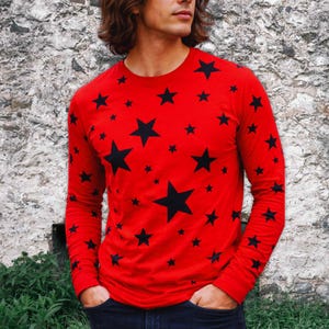 Puede incluir: Una camiseta roja de manga larga con un estampado de estrellas negras de varios tamaños. La camiseta tiene cuello redondo y está hecha de un tejido suave. Las mangas llegan hasta las muñecas. Un estilo casual.