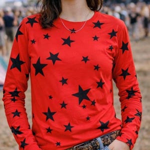 Puede incluir: Una camiseta de manga larga con cuello redondo en color rojo vibrante que presenta un patrón repetitivo de estrellas negras impresas en la parte delantera y a lo largo de ambas mangas. Se combina con vaqueros azules y un cinturón marrón.
