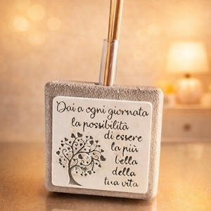 May include: A square, textured, light-colored stone diffuser base with a central clear tube holding three wooden reeds. A white rectangular plaque on the front features a black tree illustration with heart-shaped leaves and the Italian text: Dai a ogni giornata la possibilità di essere la più bella della tua vita.