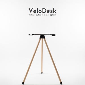 Op de afbeelding: Een VeloDesk, een draagbare bureau met een zwart tafelblad en licht houten statiefpoten. De bureau heeft een zwart blad met zijuitbreidingen en een centrale steun. De statiefpoten zijn licht hout met zwarte uiteinden. De tekst "VeloDesk" en "When outside is no option" staan erboven.