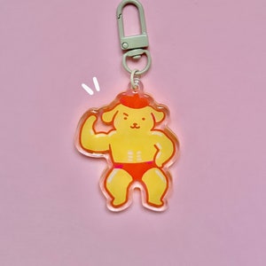 May include: A yellow dog-shaped keyring with a flexing arm, wearing an orange hat and briefs. The keyring has a silver clasp and ring. The background is a solid pink colour.