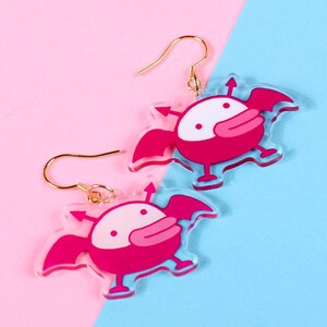 Puede incluir: Un par de pendientes de acrílico rosa con un personaje de dibujos animados con alas y un pico. Los pendientes tienen ganchos dorados.