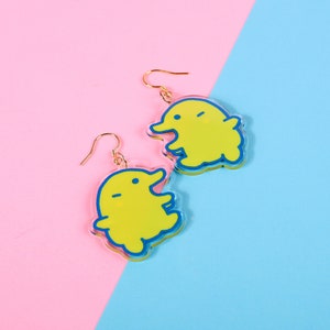 Puede incluir: Un par de pendientes amarillos colgantes con contornos azules en forma de criatura de dibujos animados. Los pendientes están sobre un fondo rosa y azul.