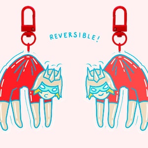 Puede incluir: Dos llaveros reversibles con un diseño de personaje de dibujos animados. Cada llavero presenta un cuerpo rojo, un contorno azul claro y un clip rojo. El personaje tiene una corona, una máscara azul y pelo rubio. El texto "REVERSIBLE!" es azul claro.