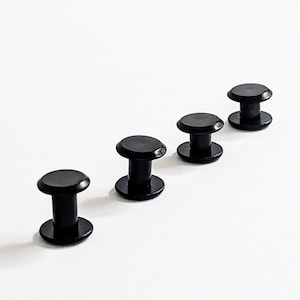 May include: Four black plastic snap fasteners, also known as press studs, are lined up in a row on a white surface.