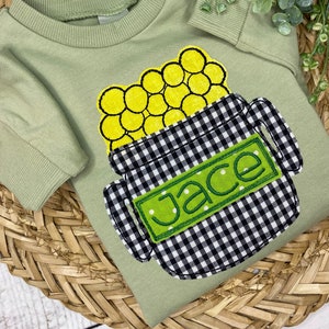 May include: A sage green short-sleeved shirt featuring an appliqué design of a black and white gingham patterned pot filled with yellow circles. A rectangular green patch with white polka dots is centred on the pot, embroidered with the name Jace.